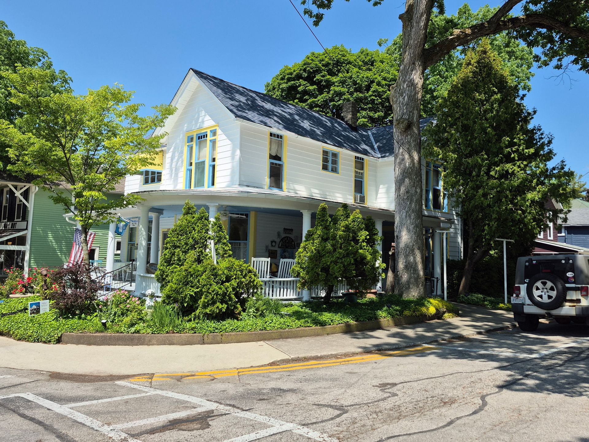 A white house with a porch and dark blue shingled roof, surrounded by green trees and shrubs on a sunny day.