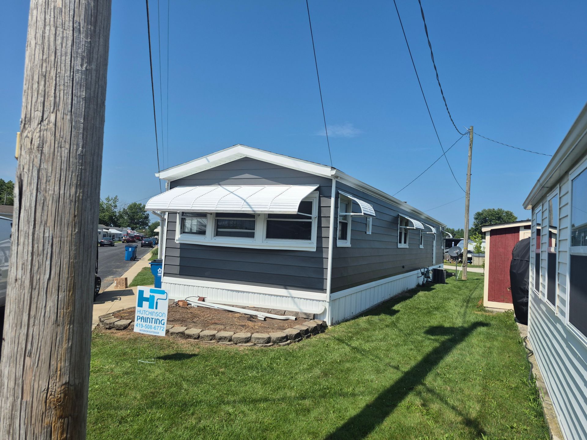 A gray, single-wide manufactured home with a white awning and a small landscaped bed in a grass yard under a blue sky.