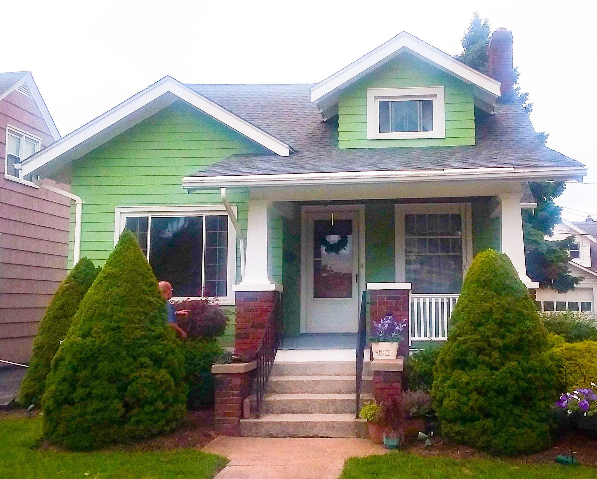 A green, one-story bungalow with a porch, brick pillars, a dormer window, and two cone-shaped evergreen bushes out front.