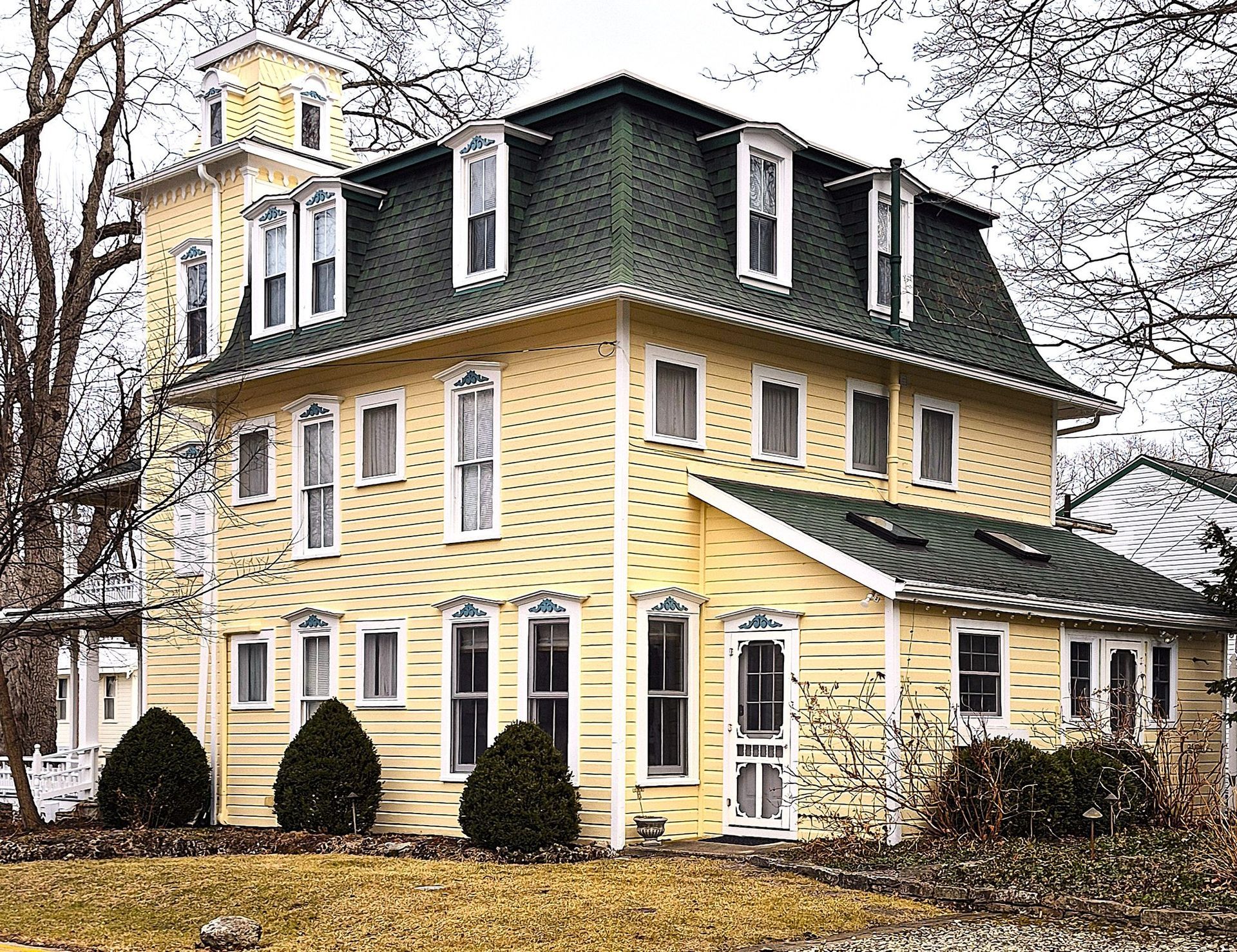 A yellow, three-story historic Victorian house with a green mansard roof, a square tower, and white trim.