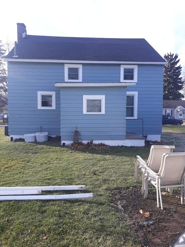 A light blue two-story house with a small center extension, a black shingled roof, and patio chairs in the grass.
