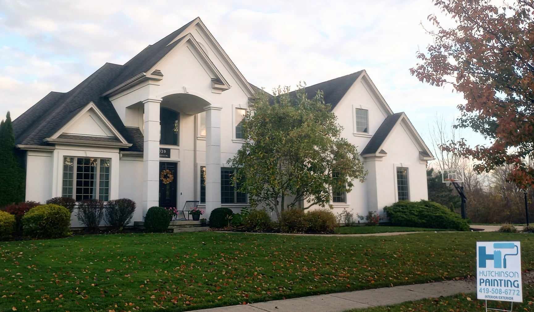 A two-story white house with dark shingles and a green lawn, featuring a front lawn sign for a painting company.