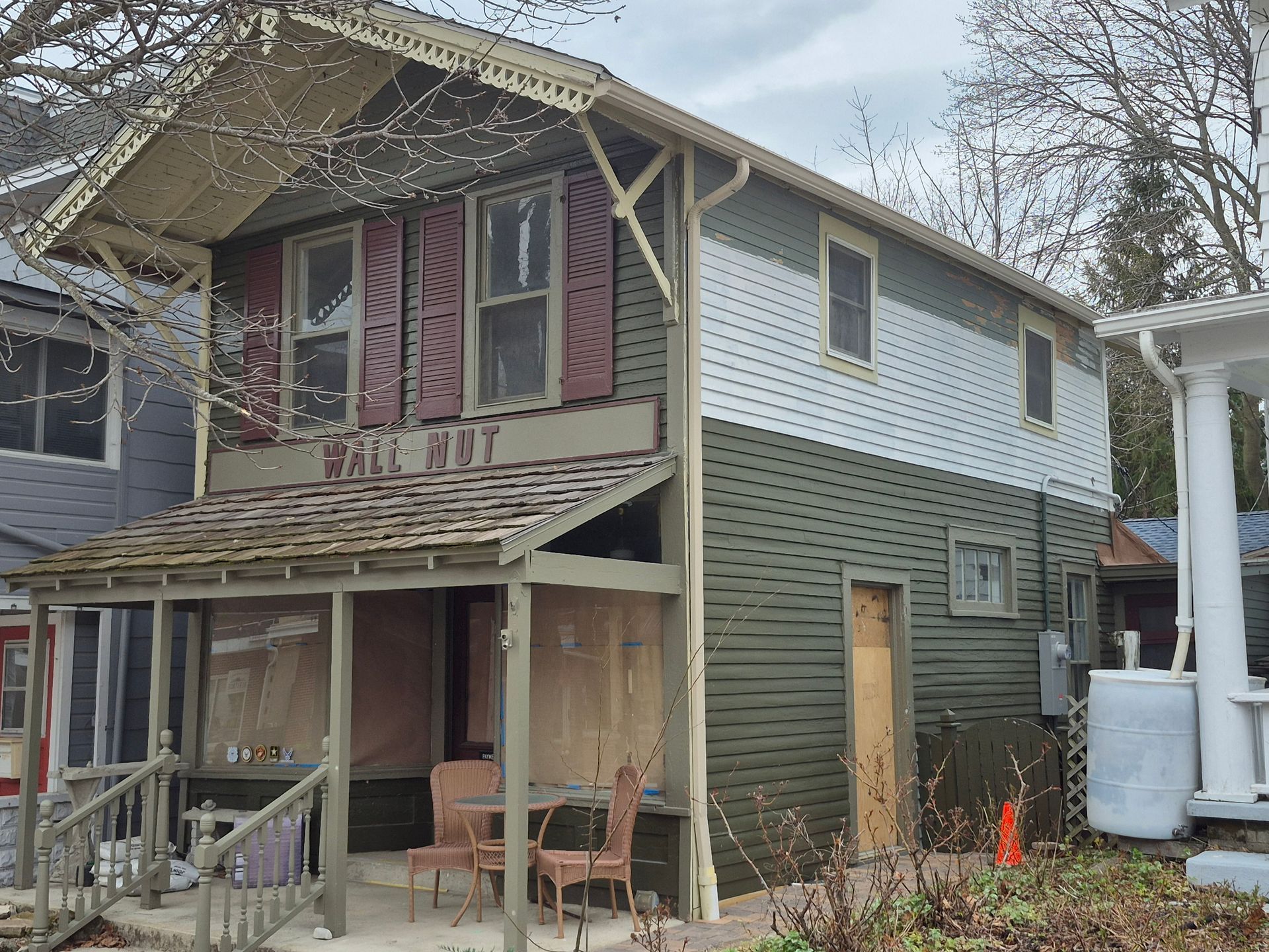 Two-story wooden house with green and white siding, a front porch, and boarded-up windows, located in a neighborhood.