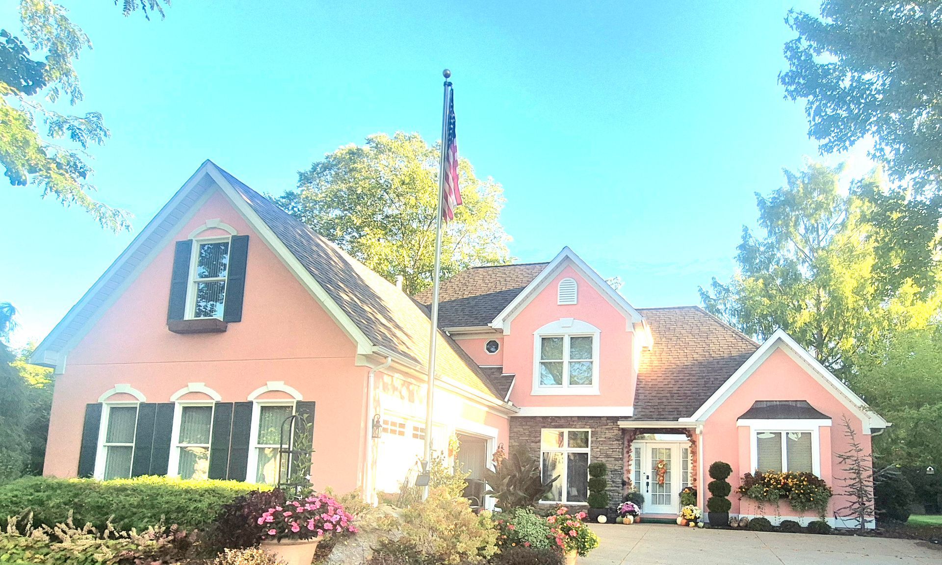 A two-story, pastel-pink house with dark shutters, a flag pole in the front yard, and landscaped gardens.