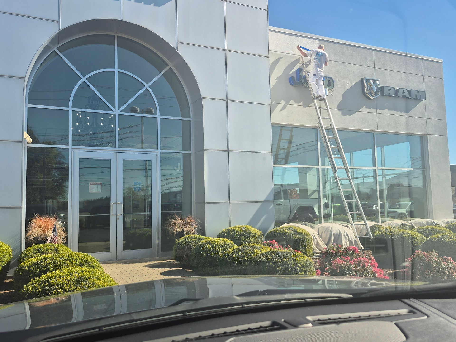 A person on a ladder works on the Ram dealership building signage above large glass windows near a glass-paneled entry.