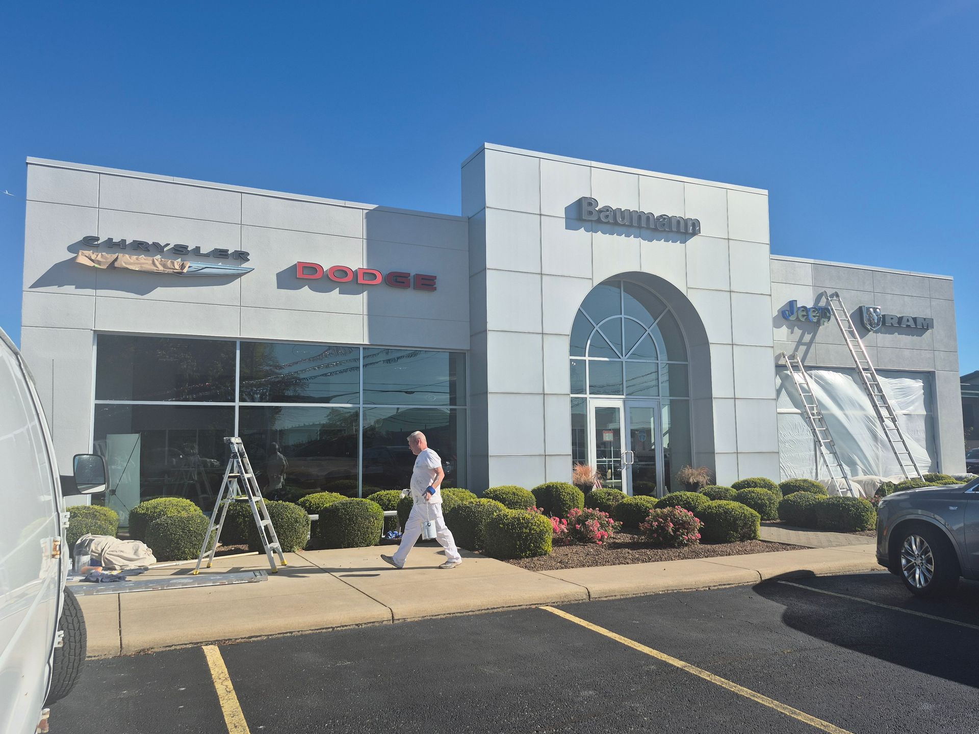 A light-colored automotive dealership building under a clear blue sky, with a worker on a ladder and another walking by.