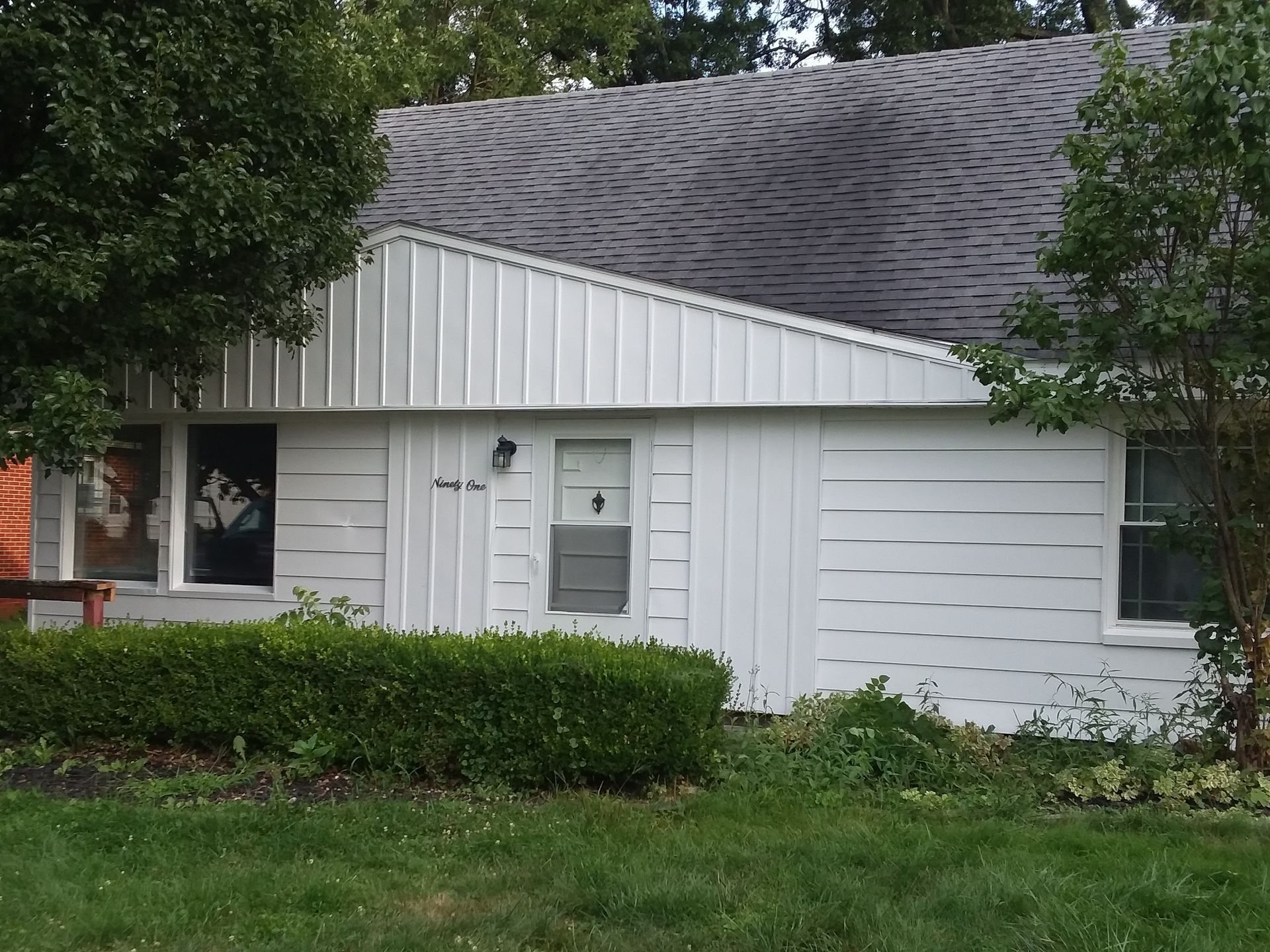 A white house with a gray shingled roof, boarded windows, and vertical and horizontal siding, partially hidden by trees.
