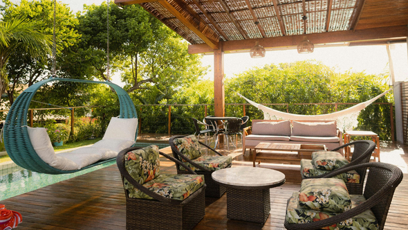 Outdoor patio with hanging chair, seating area, and hammock under a wooden pergola, surrounded by lush greenery.