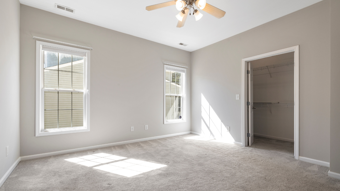 Empty bedroom with neutral gray walls, carpet, and two windows. A closet is in the right corner.