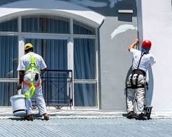 Two painters on a roof, one holding a bucket, the other painting a white building wall.