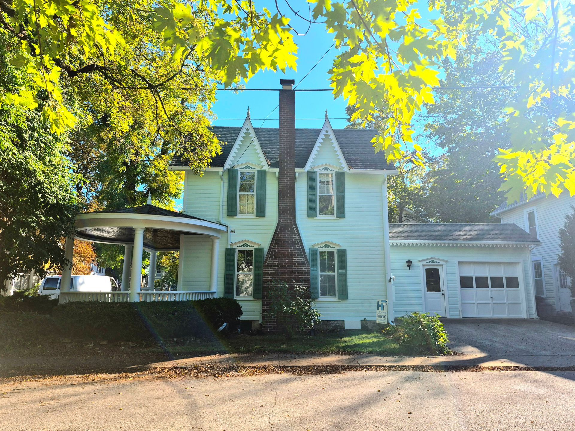 A light green two-story house with a round porch, brick chimney, and attached garage surrounded by autumn trees.