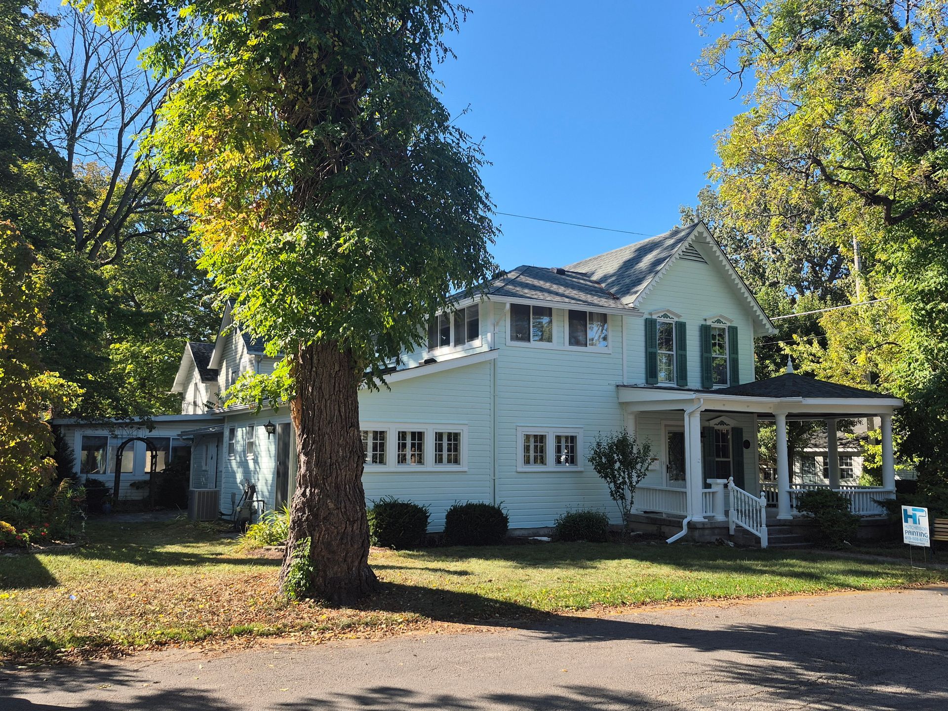 A light-green two-story Victorian house with a front porch, surrounded by large, leafy trees on a sunny day.