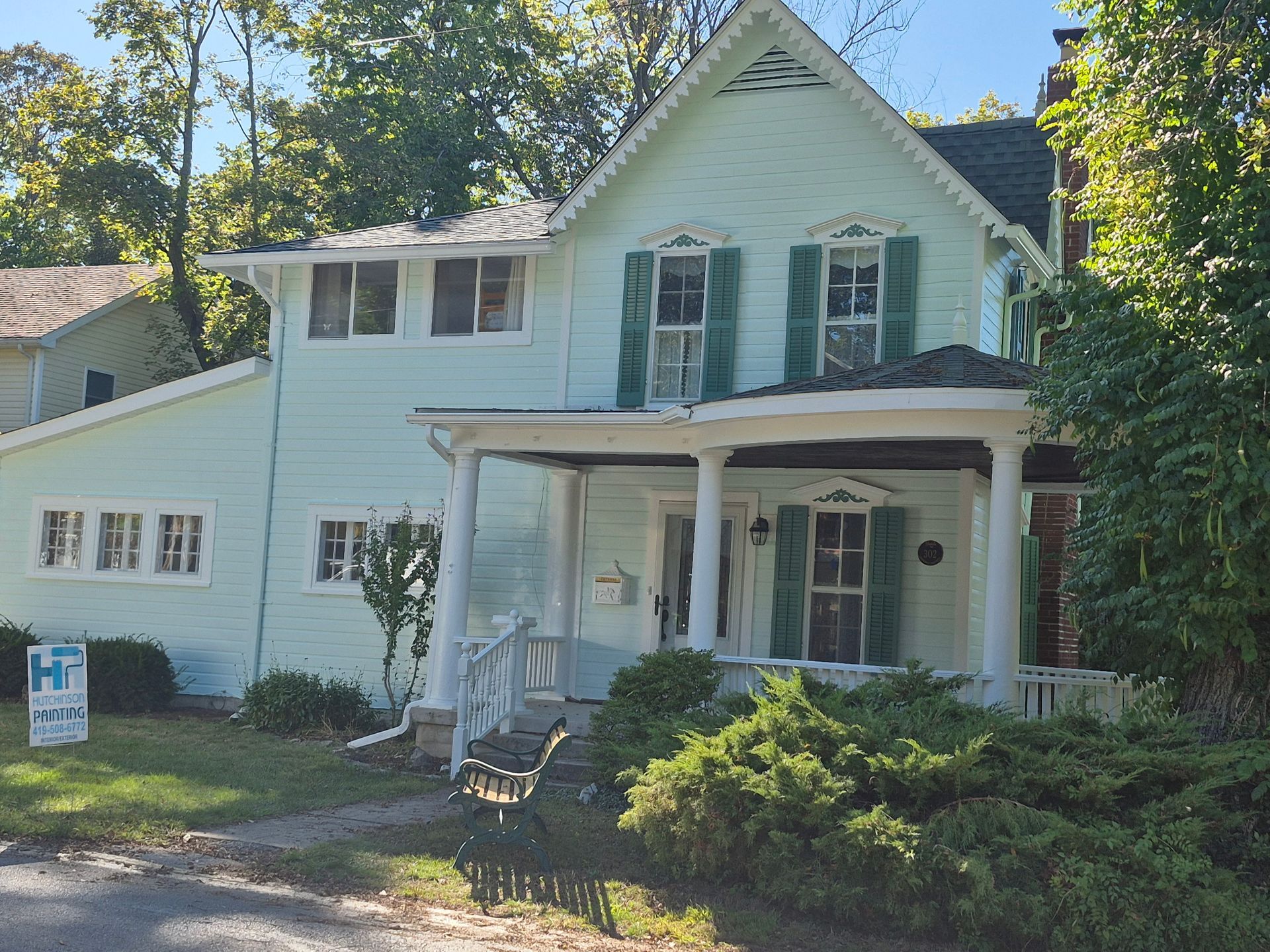 A light green, two-story Victorian house with a front porch, green shutters, and surrounding trees.