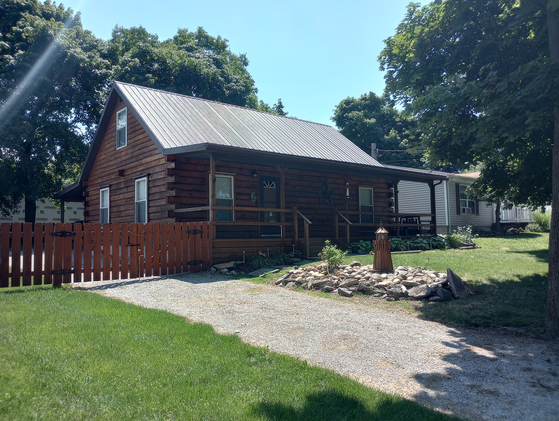A dark brown log cabin with a silver metal roof, a gravel driveway, and a red wooden fence on a sunny day.