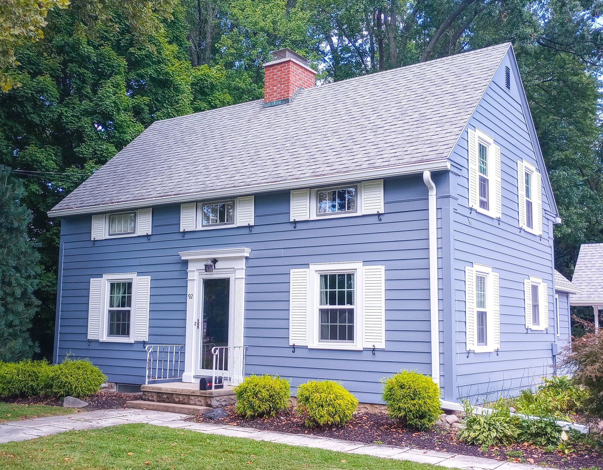 A two-story blue house with white shutters, a centered front door, and a brick chimney, surrounded by lush green foliage.