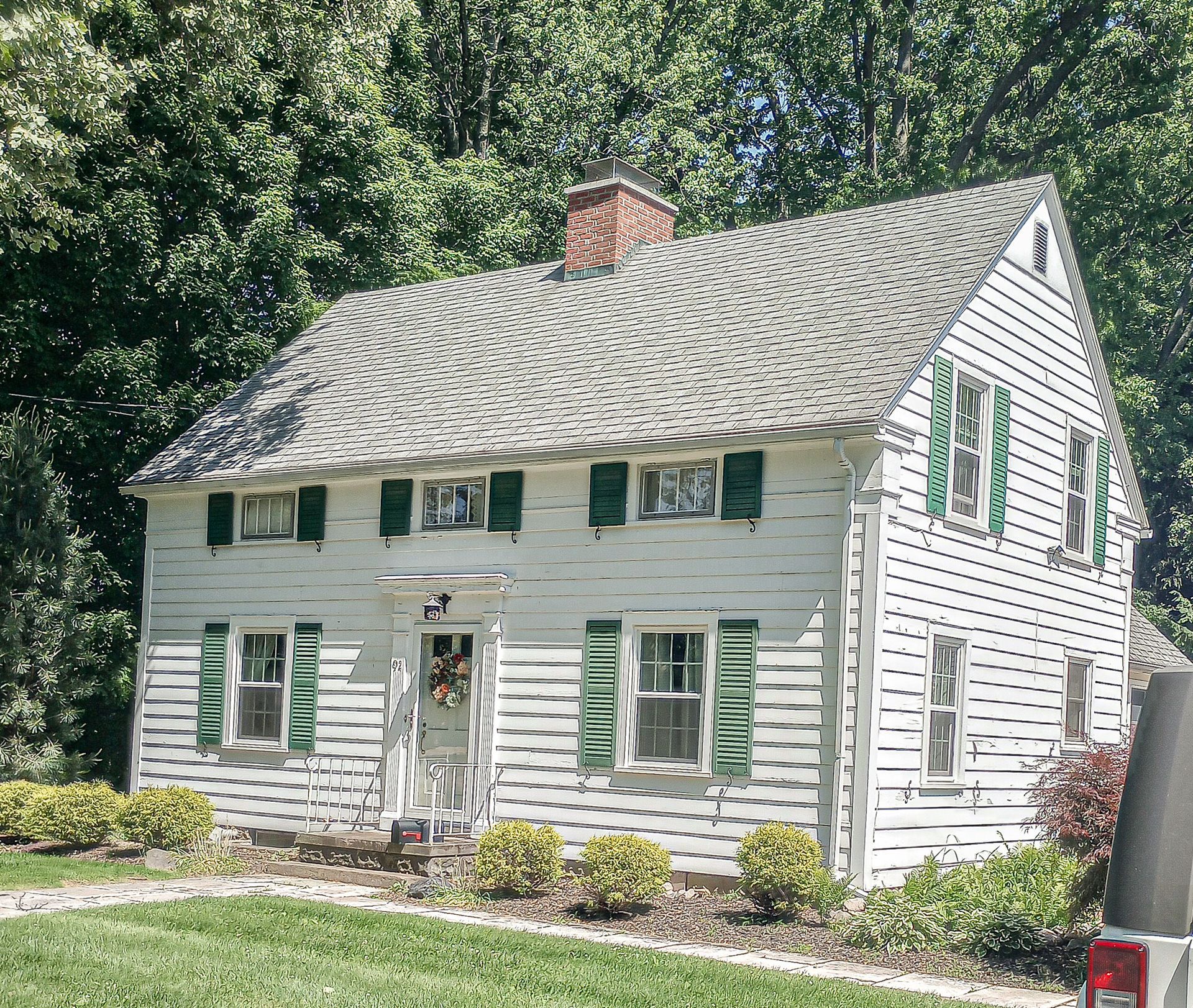 A two-story white colonial-style home with green shutters, a brick chimney, and surrounding trees and bushes.