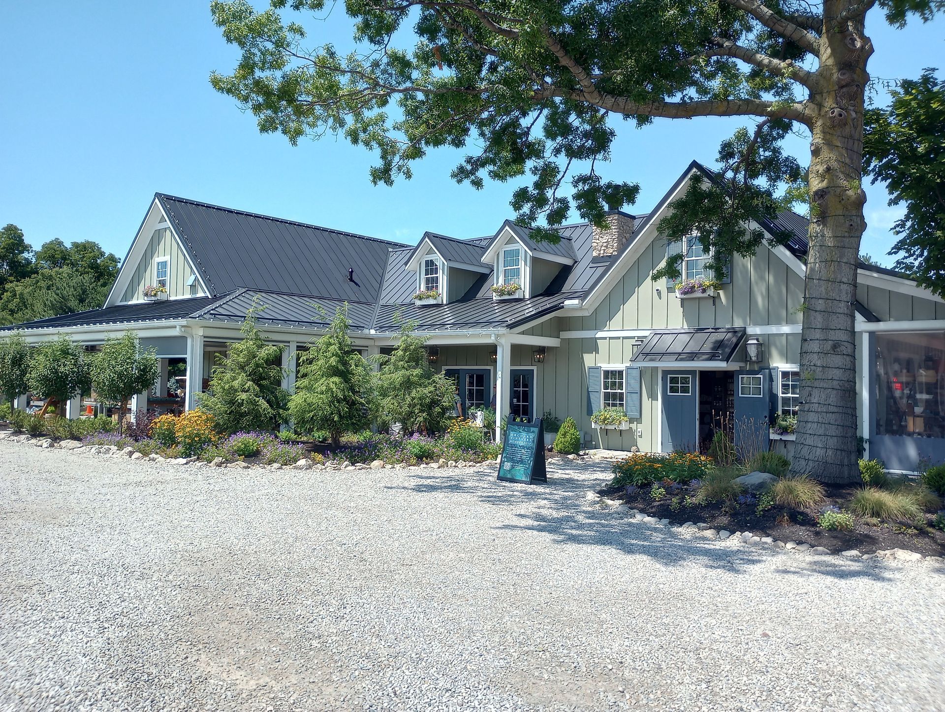 A light-green house with a dark roof and a covered porch, surrounded by trees and a gravel driveway on a sunny day.