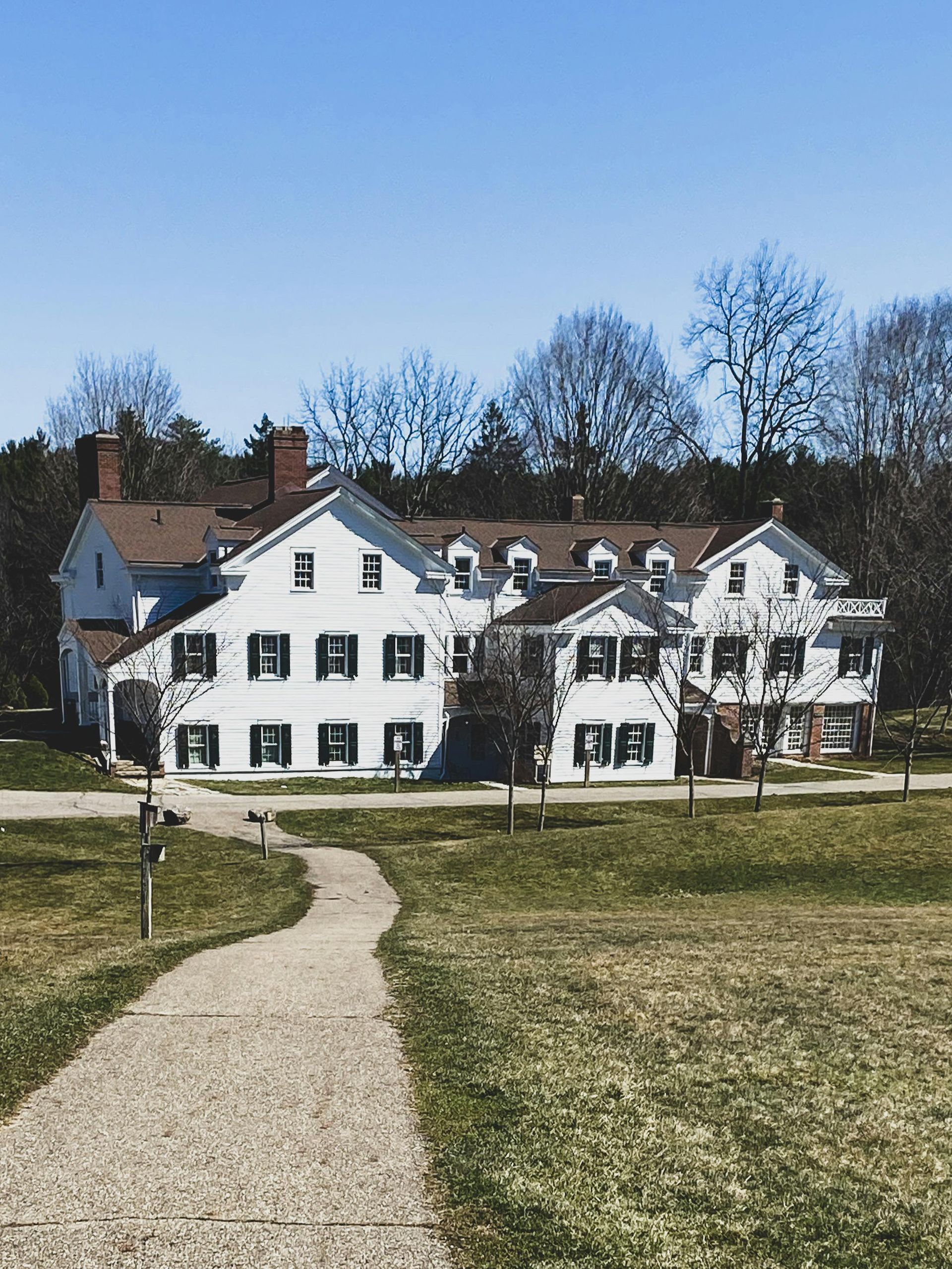 White house with black shutters, brown roof, and a gravel path leading to it, sunny day.
