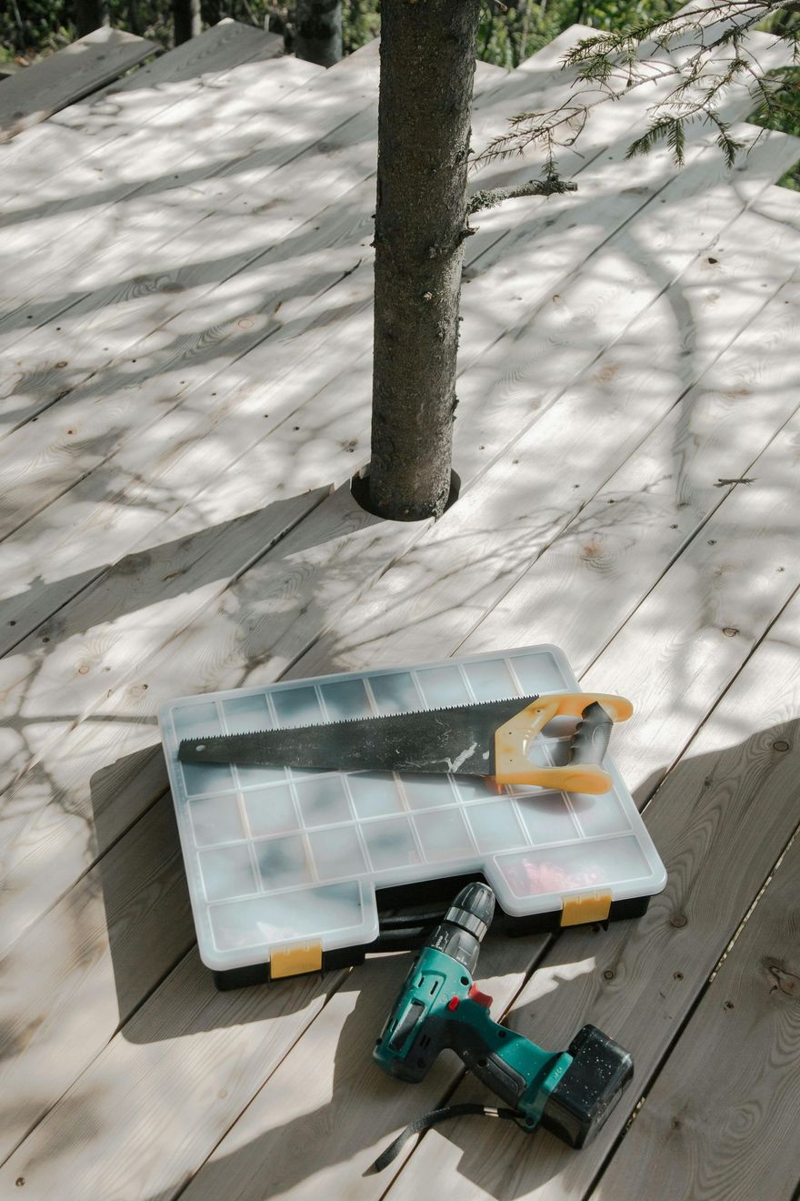 Tools on wooden deck: saw, drill, and tool box next to tree trunk with surrounding sunlight.