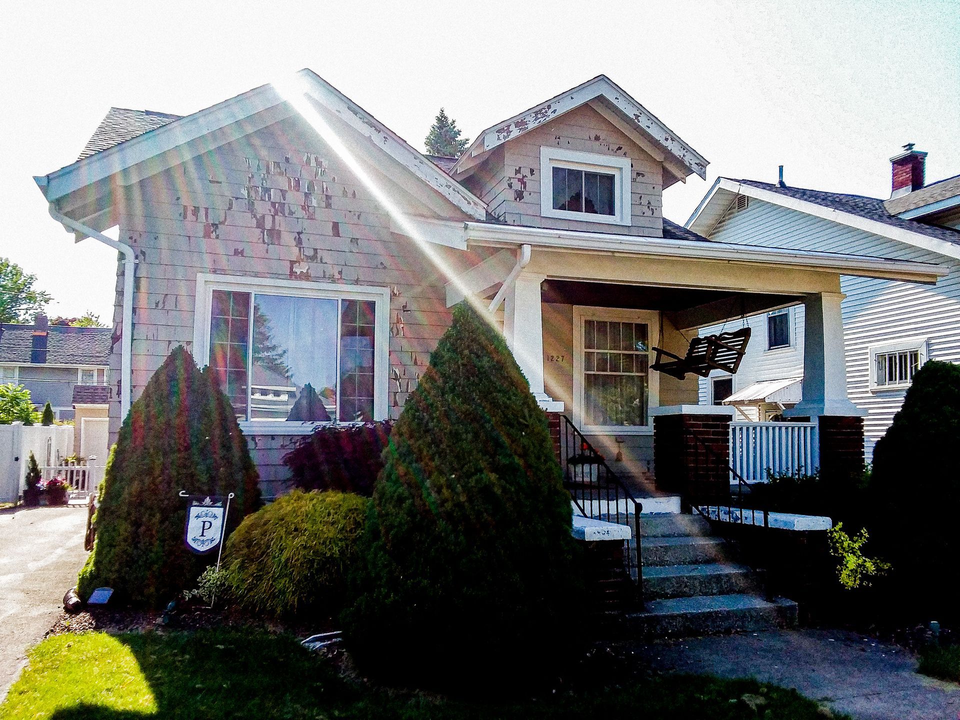 A light-colored house with a brick facade, large front window, and a covered porch, set back behind two prominent conifers.