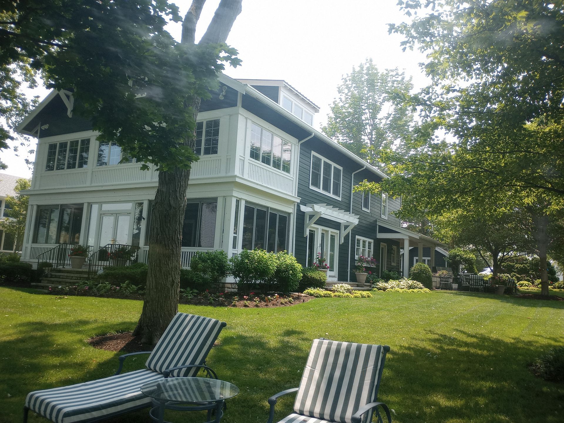 A large, two-story house with dark gray siding and white trim sits behind two striped lounge chairs on a grassy lawn.