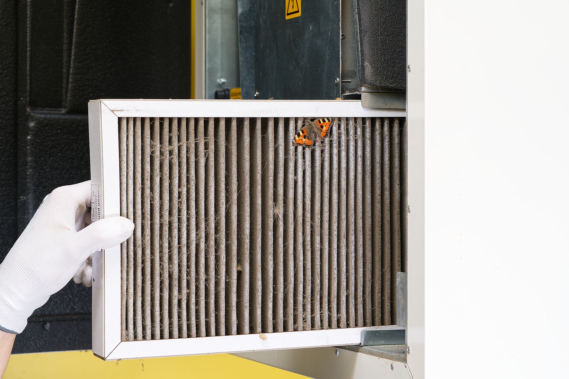 A person wearing a white glove pulls a dirty, dust-covered HVAC air filter from a unit.