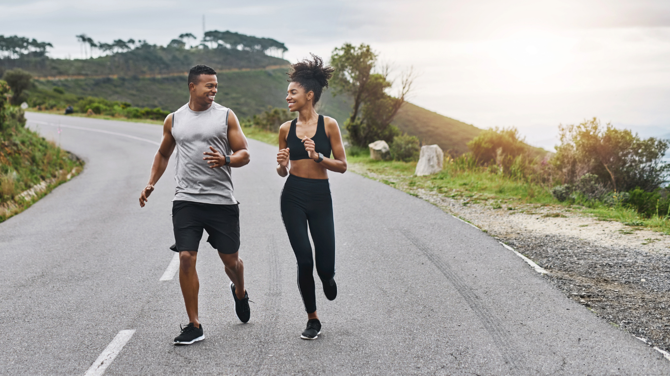 Couple running on a paved road next to a hill, smiling. Bright sunlight.