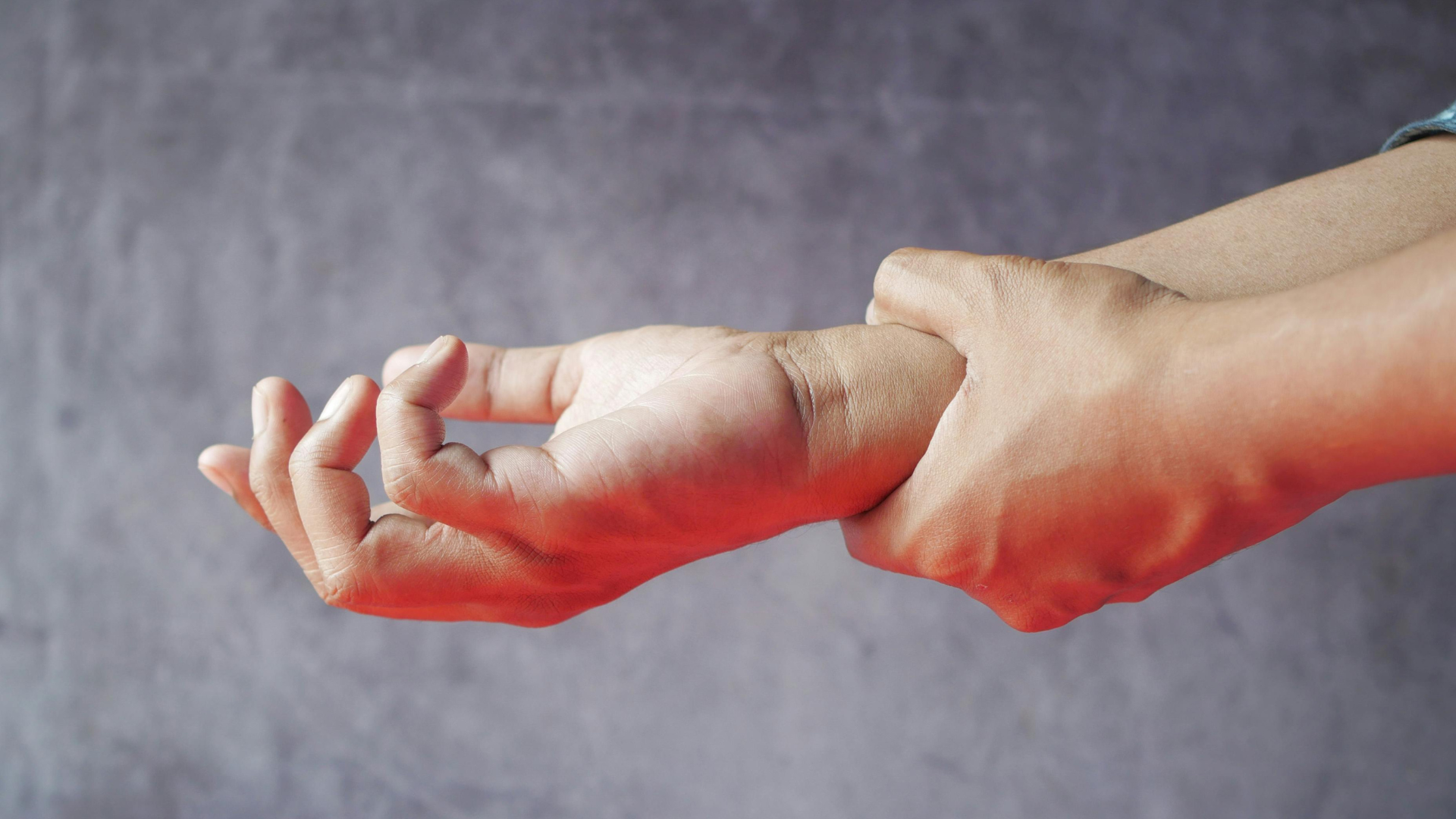 Person holding their wrist, showing signs of pain or injury, with a neutral background.