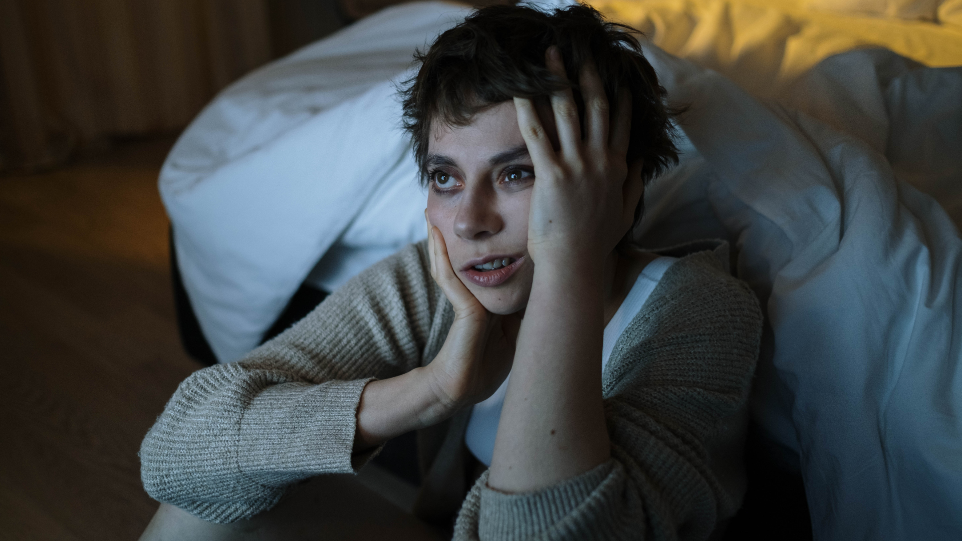 Woman in distress sits on floor next to bed, hands on head, looking worried. Dimly lit room.