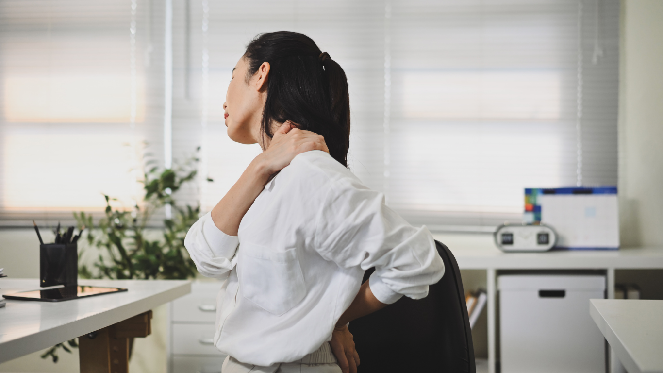 Woman in office, hand on neck and back, experiencing pain while seated.