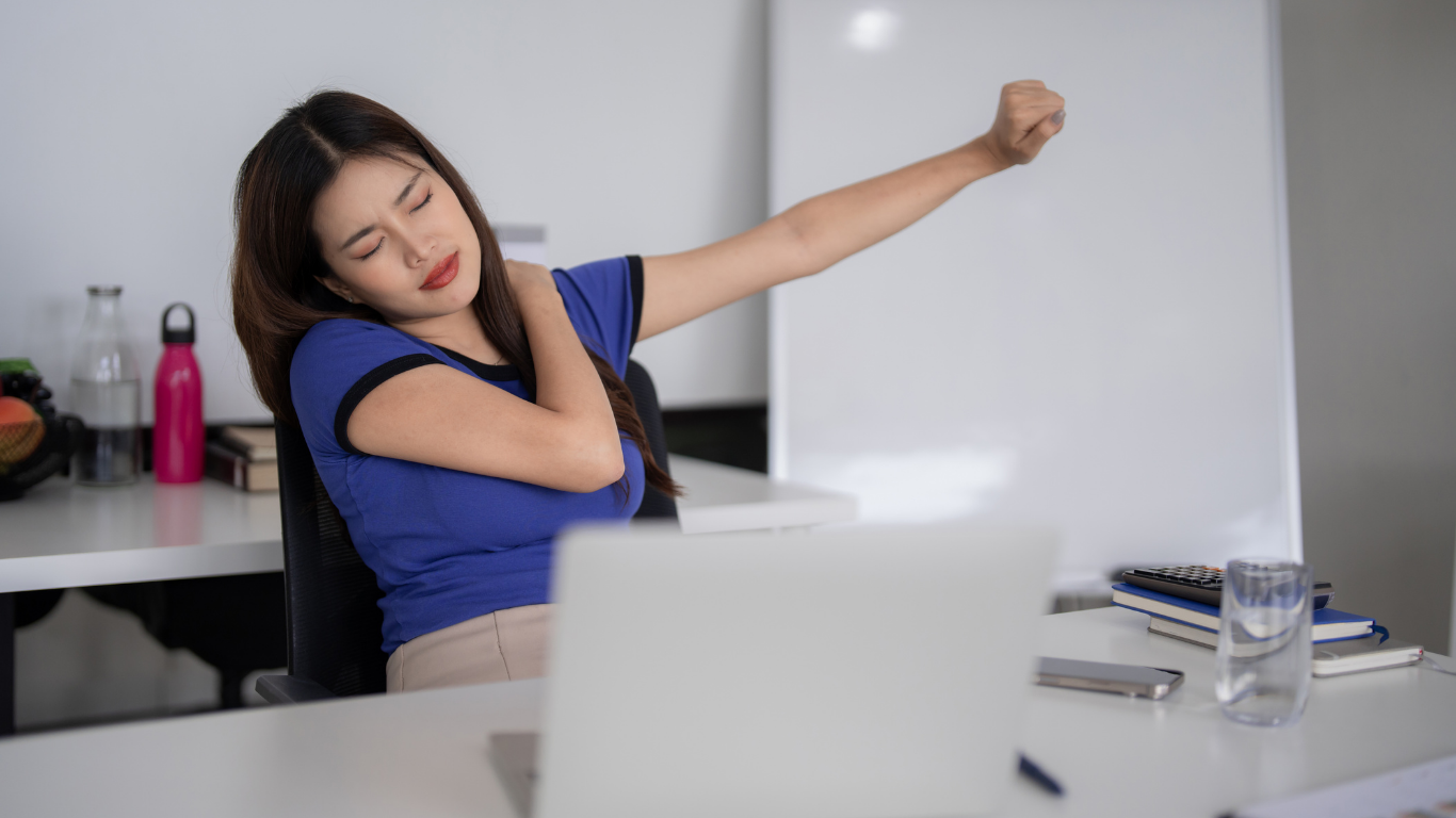 Woman in blue shirt stretching arm, sitting at desk, appears to have shoulder pain.