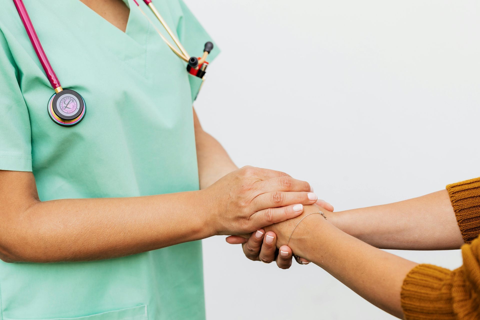 Nurse in green scrubs holding a patient's hand for comfort, stethoscope visible.