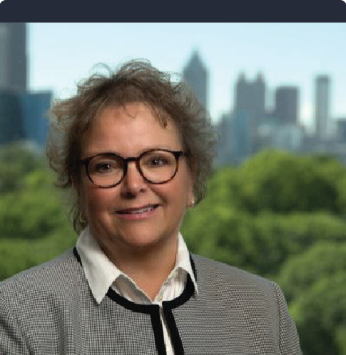 A woman wearing glasses and a plaid jacket is smiling in front of a city skyline.
