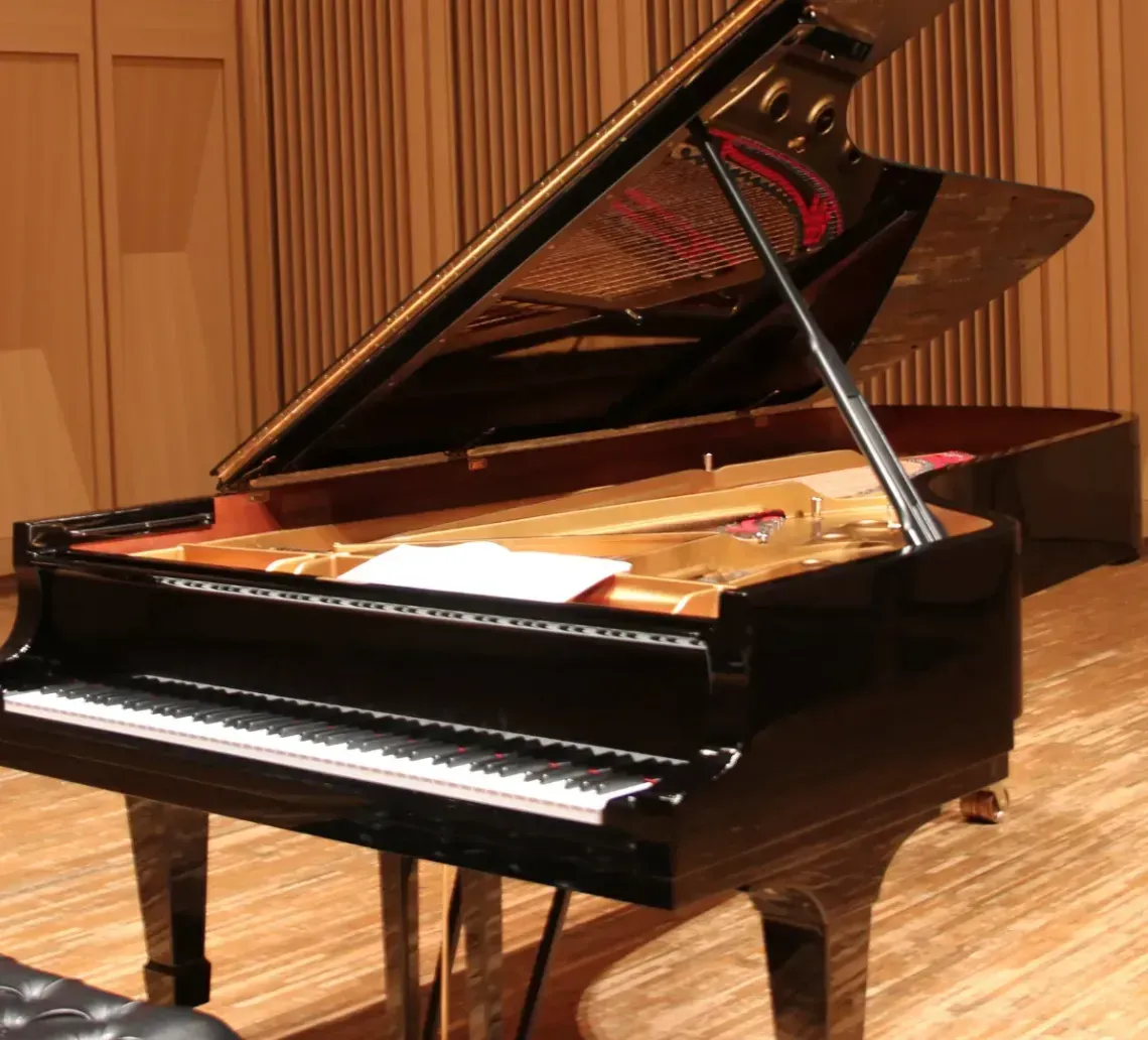A glossy black grand piano with a music sheet on the strings, centered in a room with light wood walls and flooring.