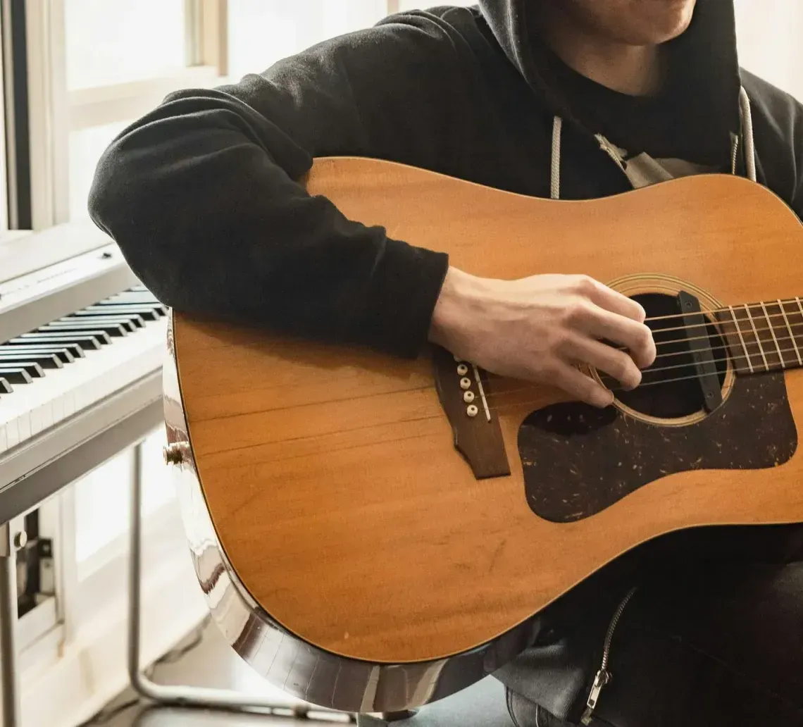 A person in a black hoodie plays an acoustic guitar next to a white keyboard.