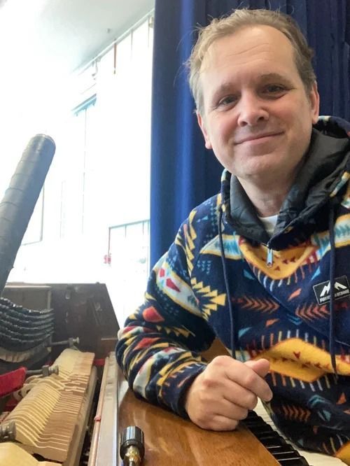 A person smiling while working on the internal mechanisms of an upright piano in a room with blue curtains.