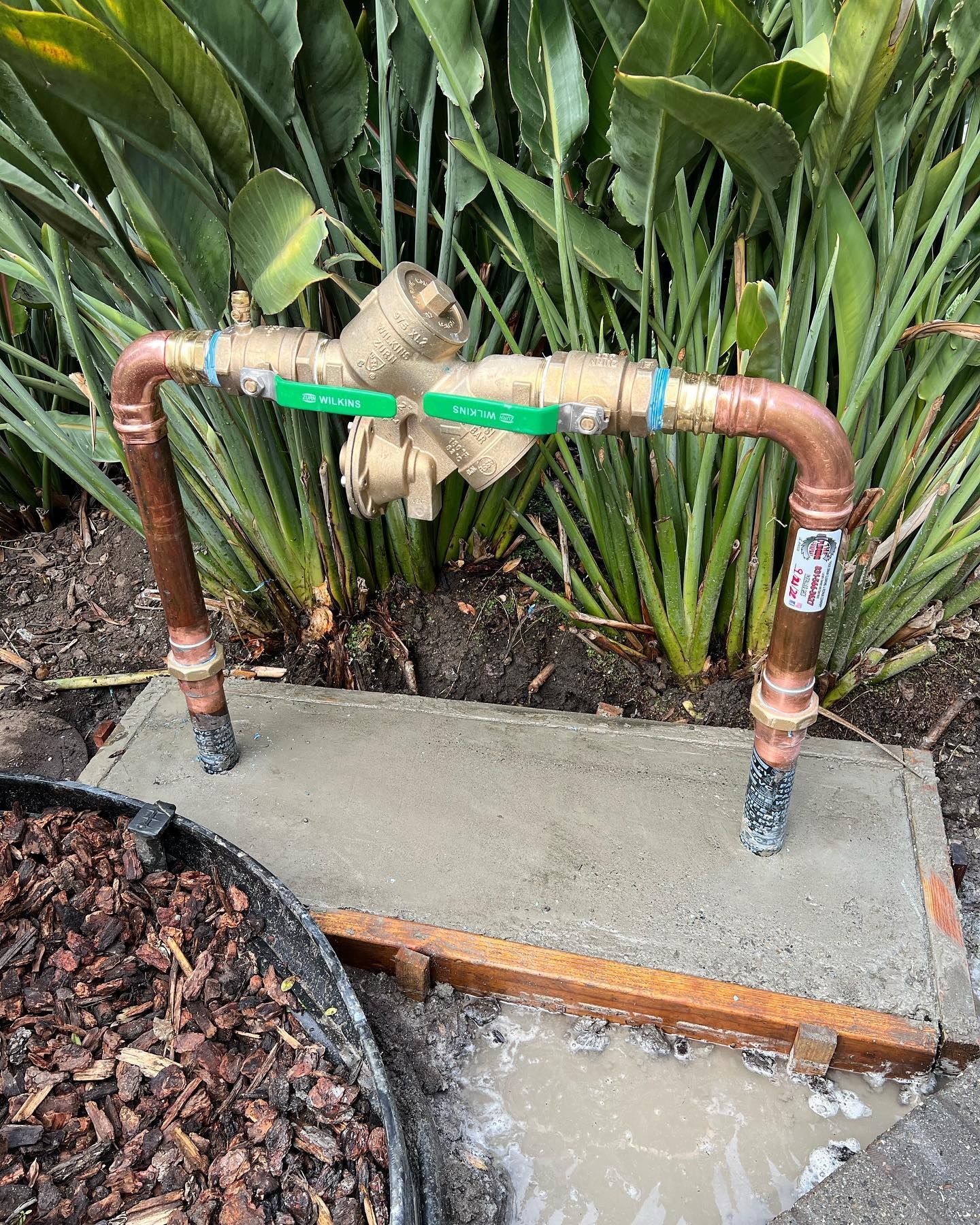 Copper and brass irrigation valve system on concrete pad. Green handles and copper piping. Plants in background.