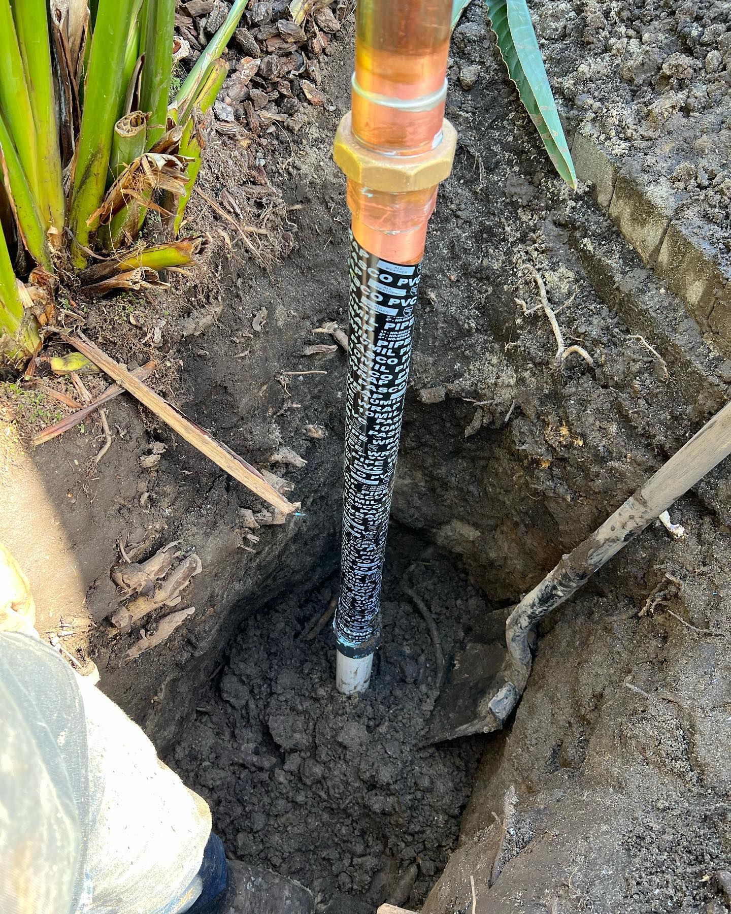 Copper pipe emerging from a hole in the dirt, connected to a brass fitting. A shovel lies nearby.