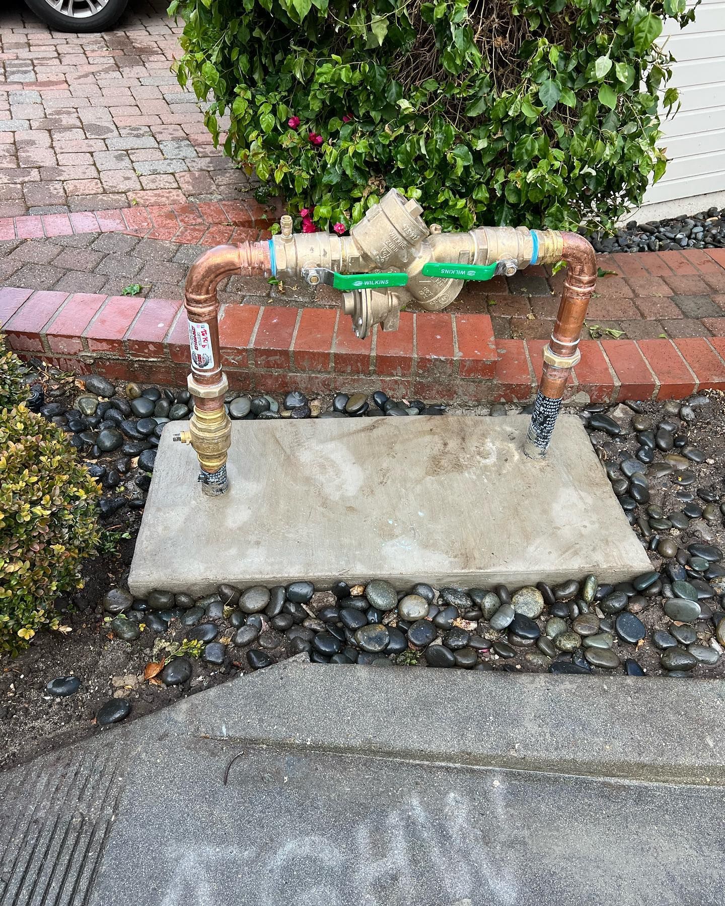 Water meter setup with copper pipes, green valve, on concrete slab, surrounded by black rocks and brick.