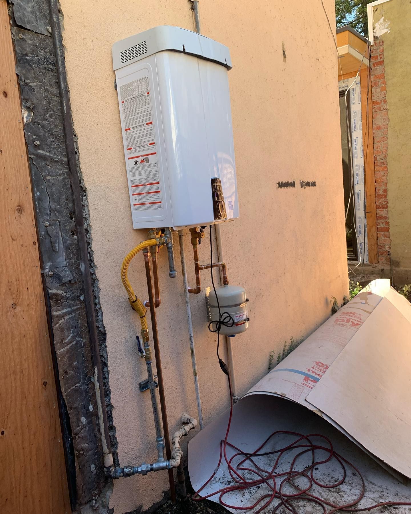 White tankless water heater mounted on a tan exterior wall with copper pipes and yellow gas line.