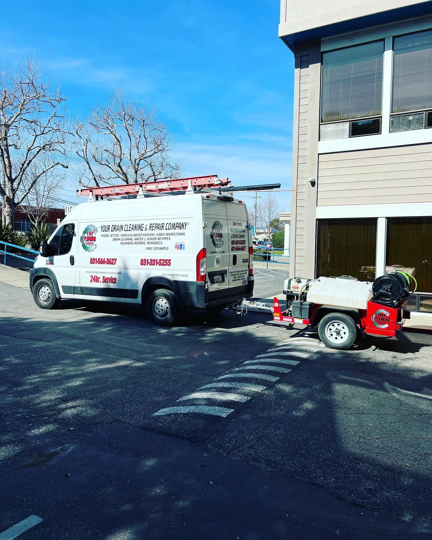 White work van and trailer parked near a building, under a blue sky. Van has company logo.