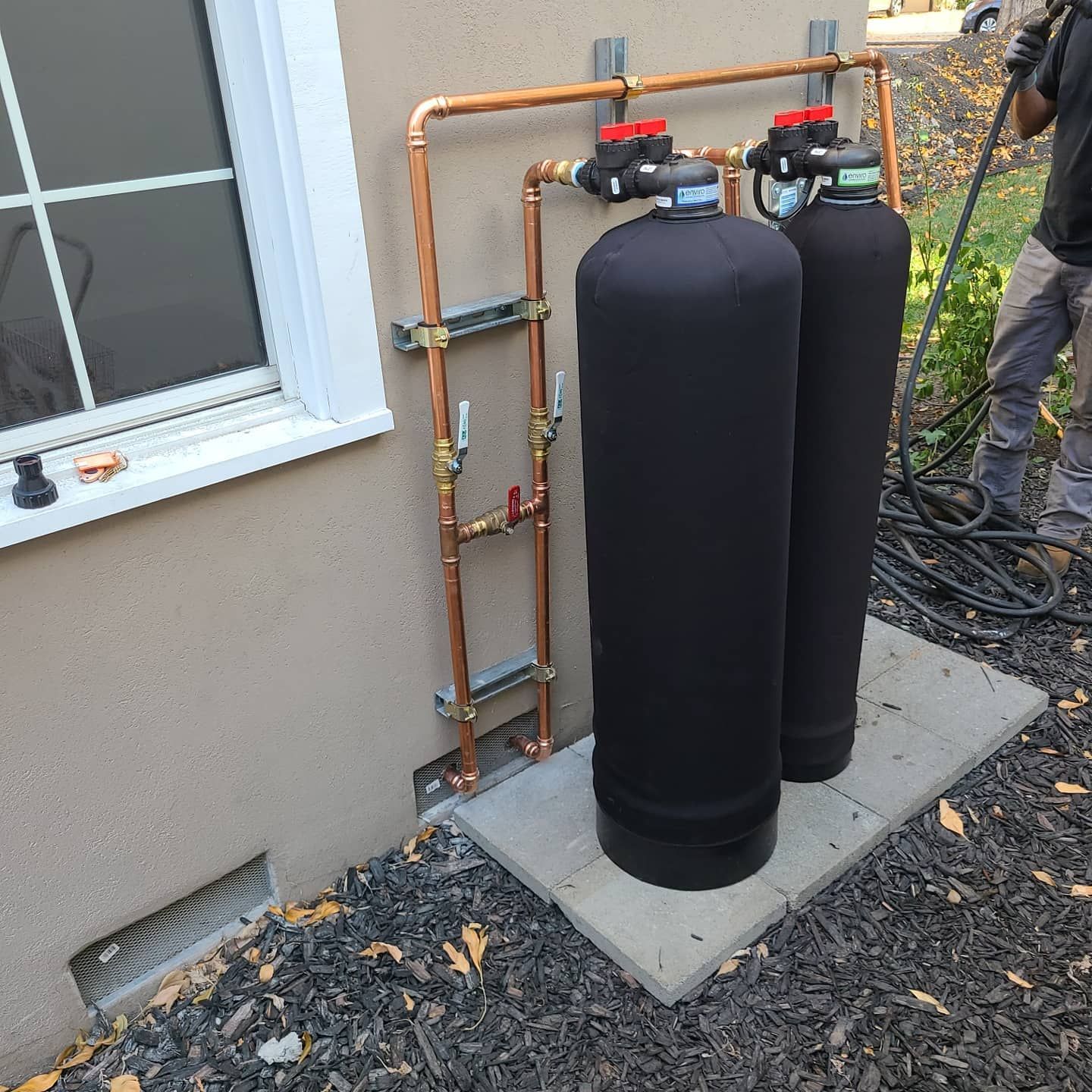 Water treatment system with copper pipes on exterior wall. Two black tanks sit on a concrete pad.
