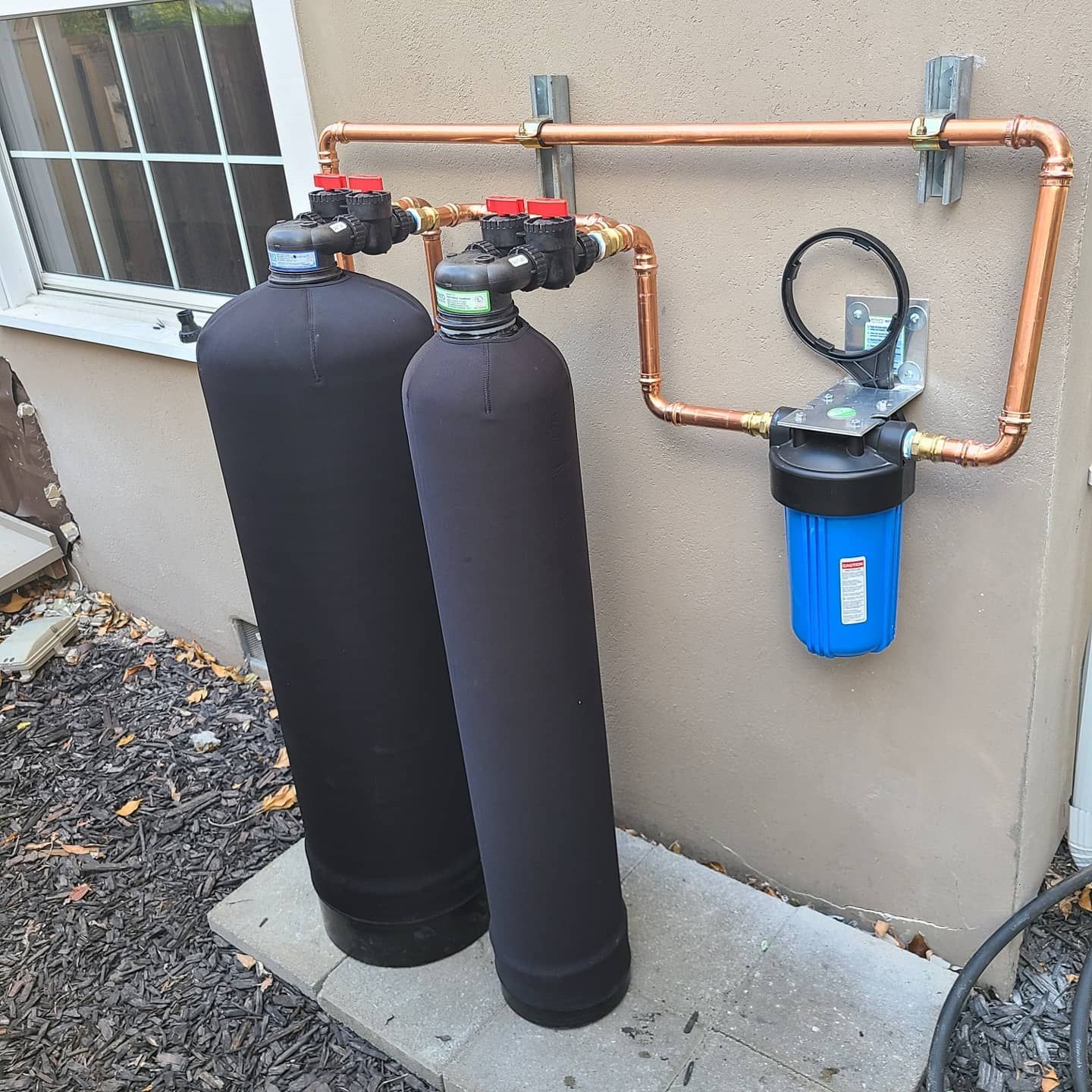 Water filtration system with two black tanks, copper piping, and a blue filter against a wall.
