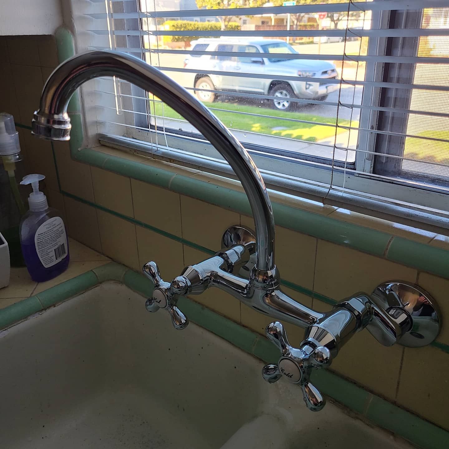 Chrome kitchen faucet with cross handles over a white sink, set against a window with a view of a car.