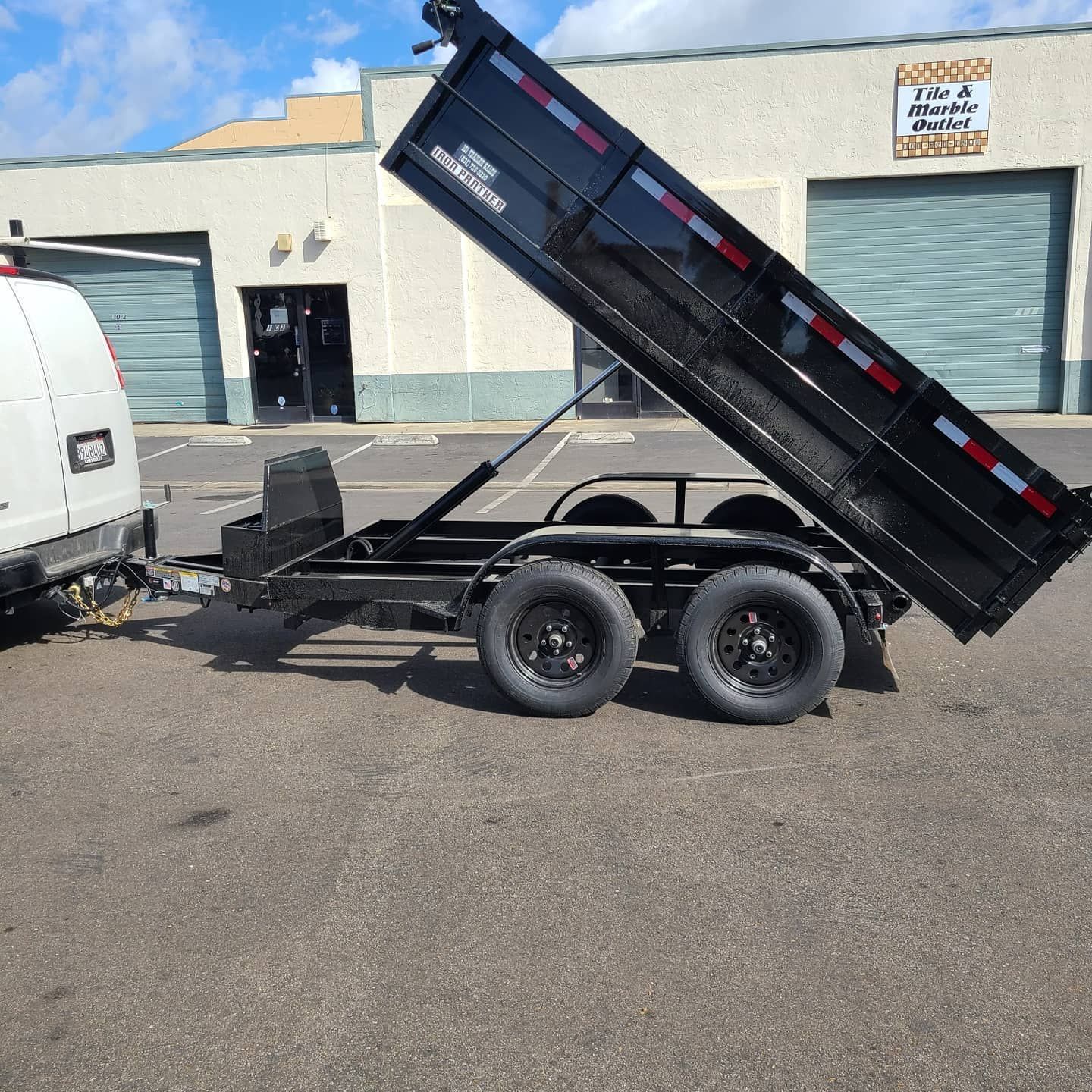 Black dump trailer hitched to a white van, tilted up; parked in front of a building.