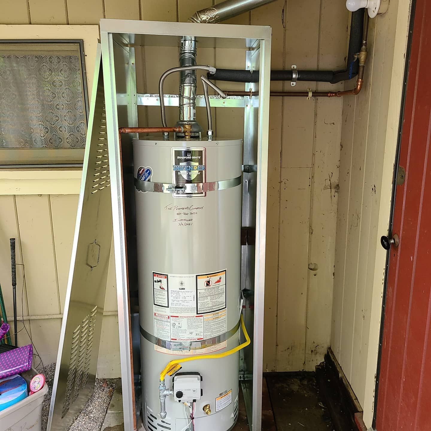 Gas water heater in a metal enclosure against a wooden wall; copper pipes visible.
