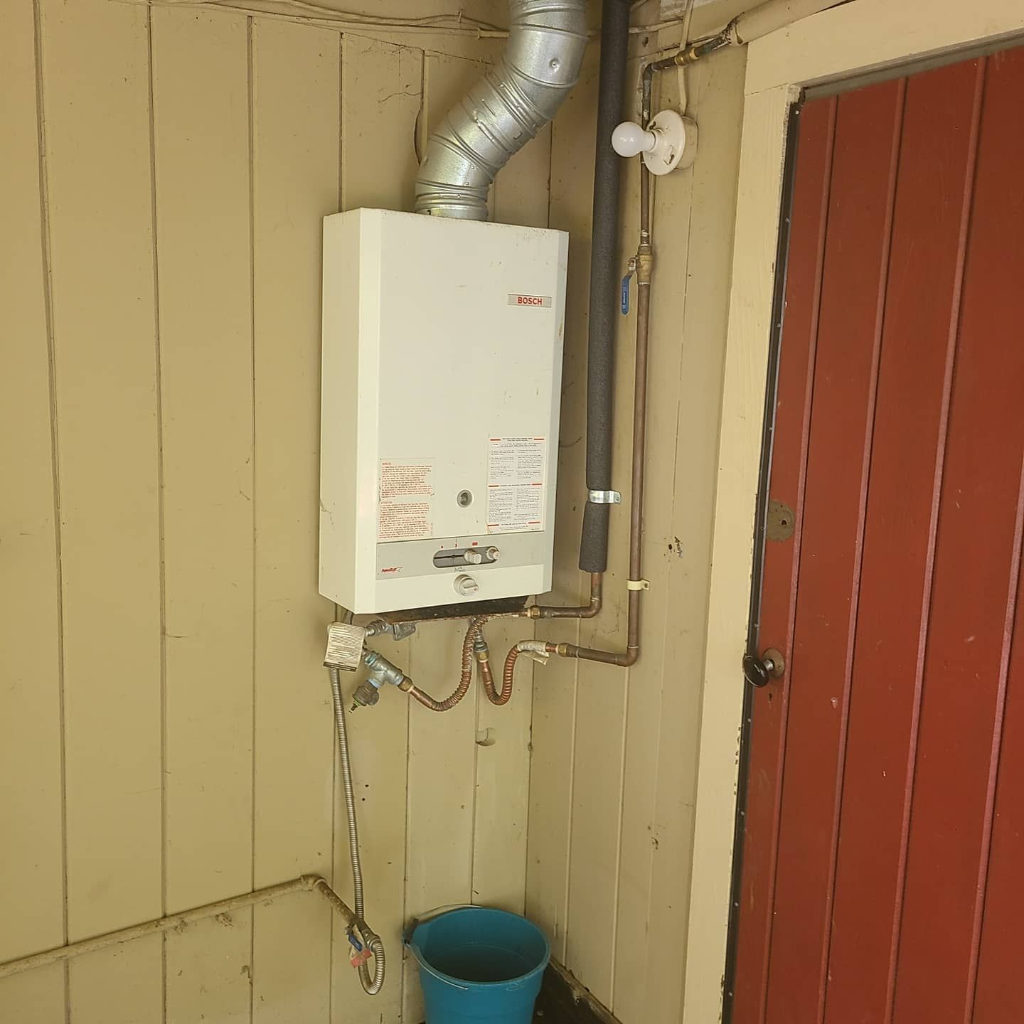 Gas water heater on a wall with copper pipes, vent, and a red door.