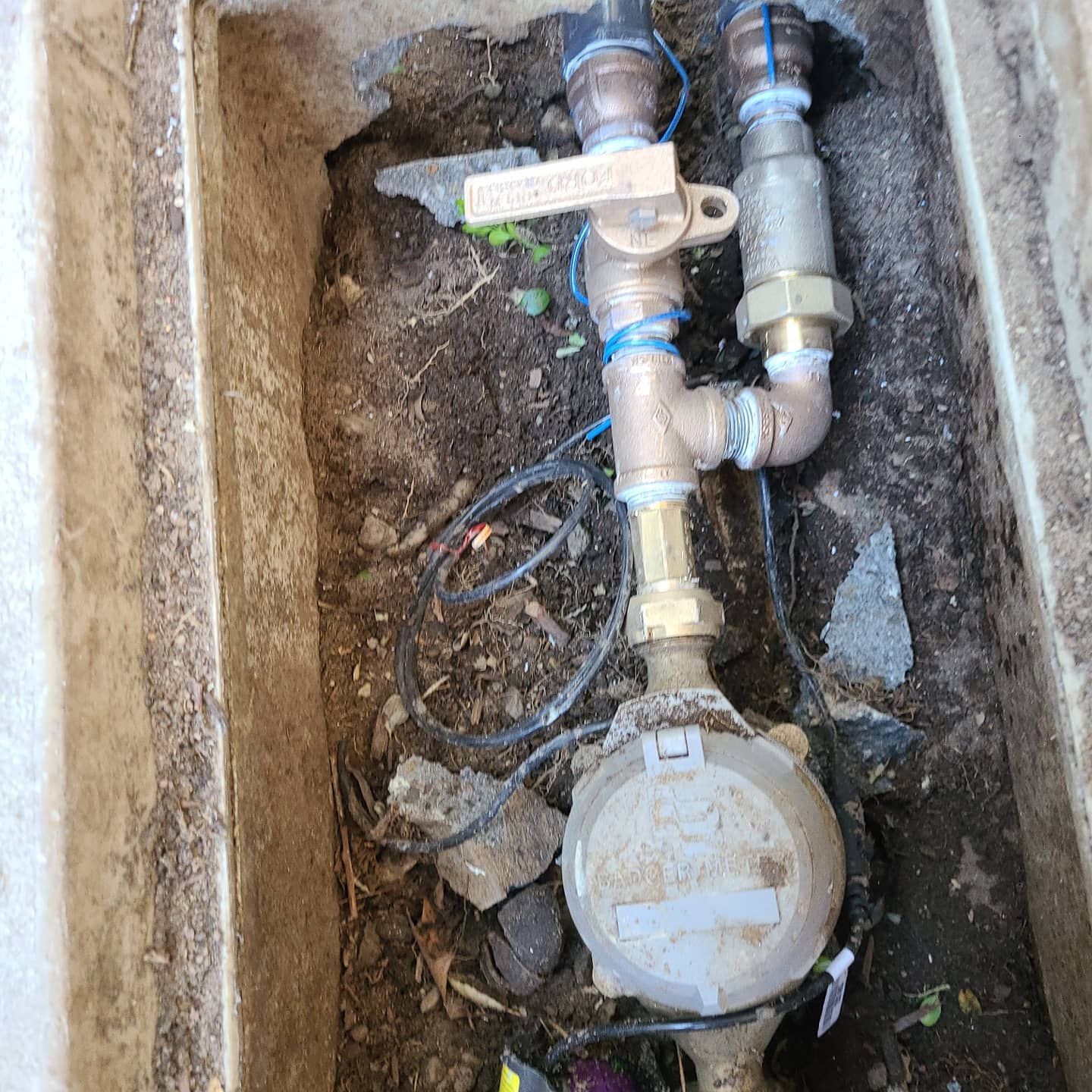 Water meter and pipes in a rectangular concrete box, surrounded by soil and debris.