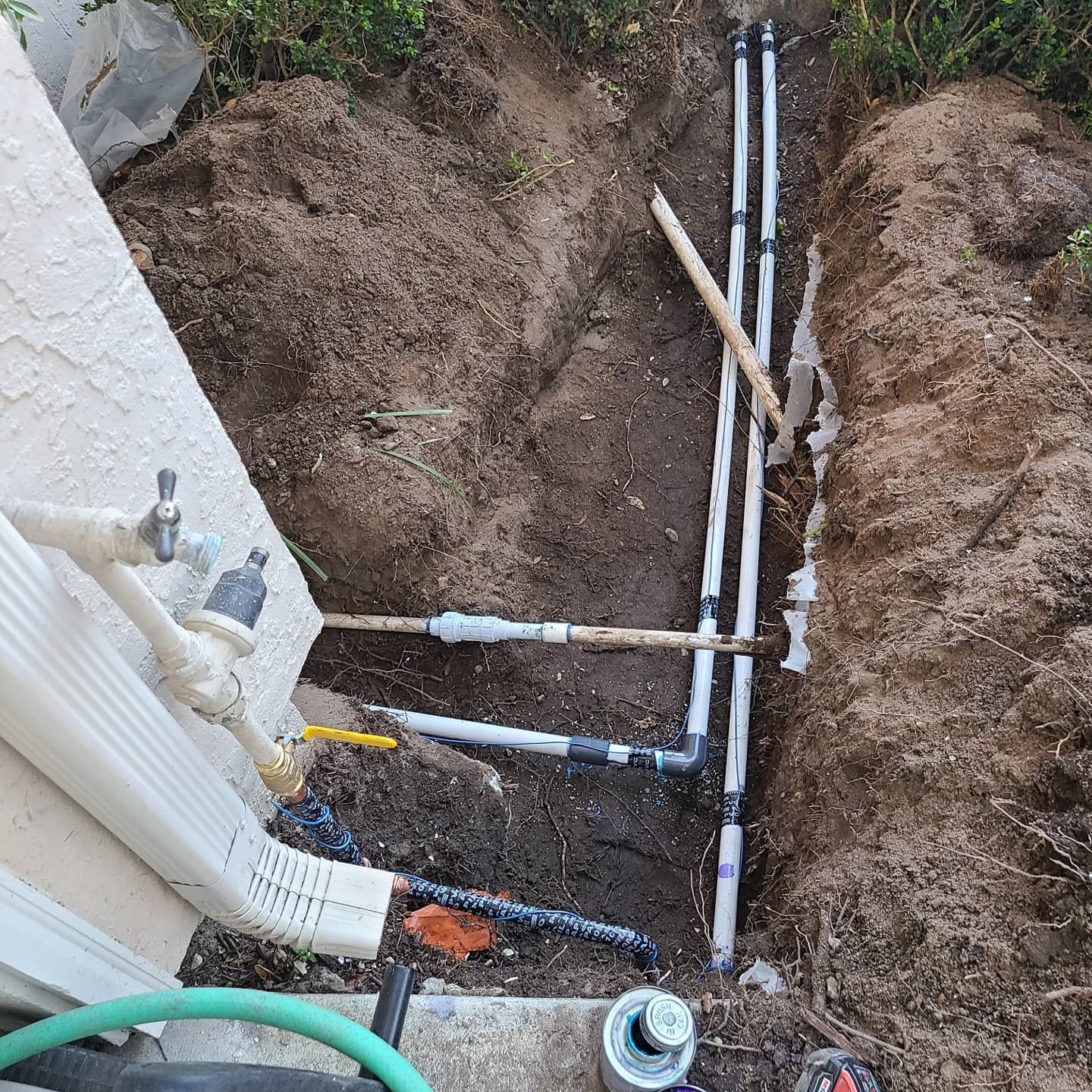 Plumbing work in progress: pipes and fittings in a trench beside a building, surrounded by dirt and grass.
