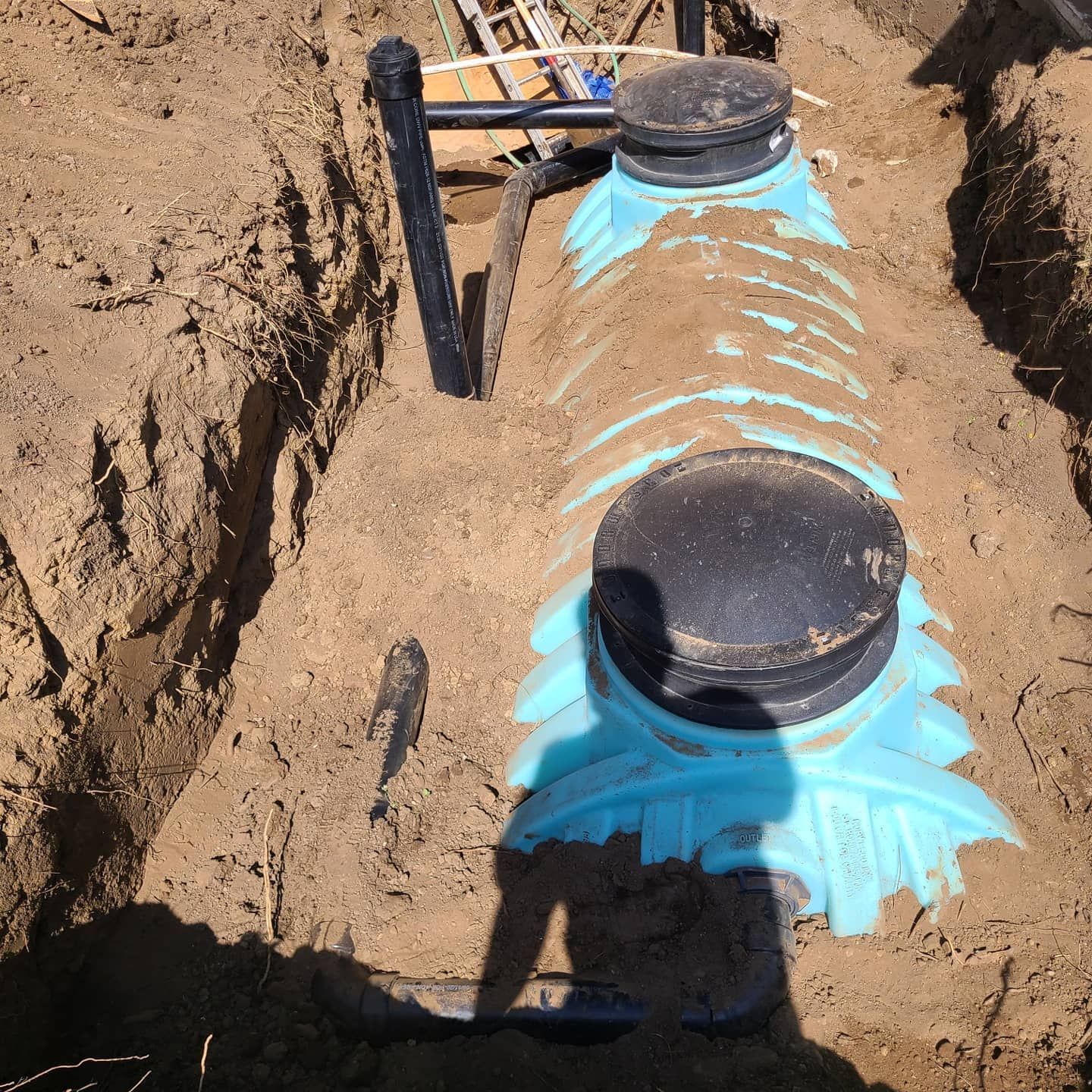 Blue plastic septic tank sections in an excavated trench, with black access lids and connected piping.